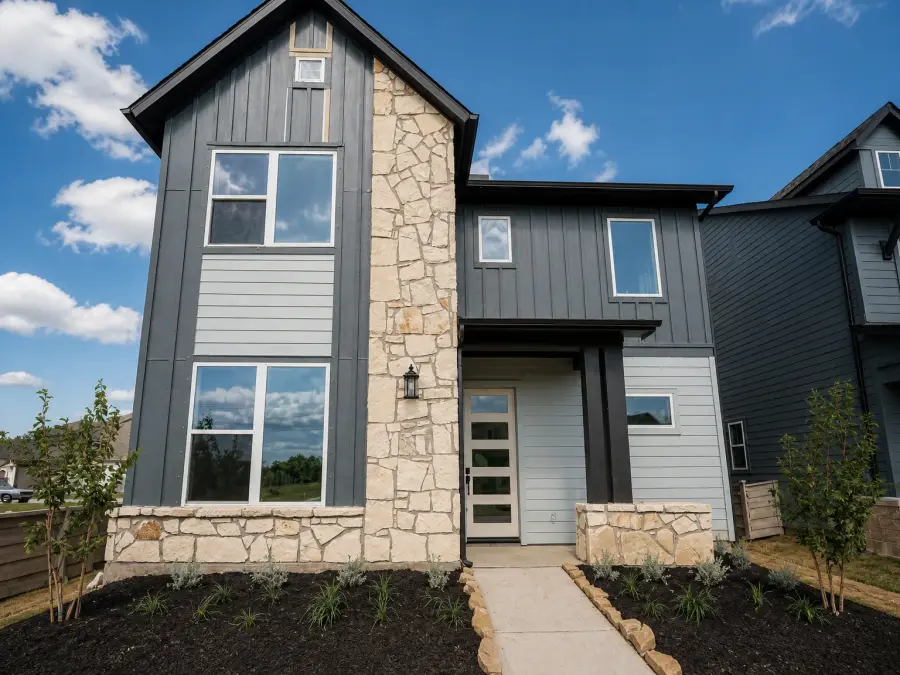 Before day to dusk conversion, a modern two-storey home with grey board-and-batten siding and a limestone chimney under a clear midday sky