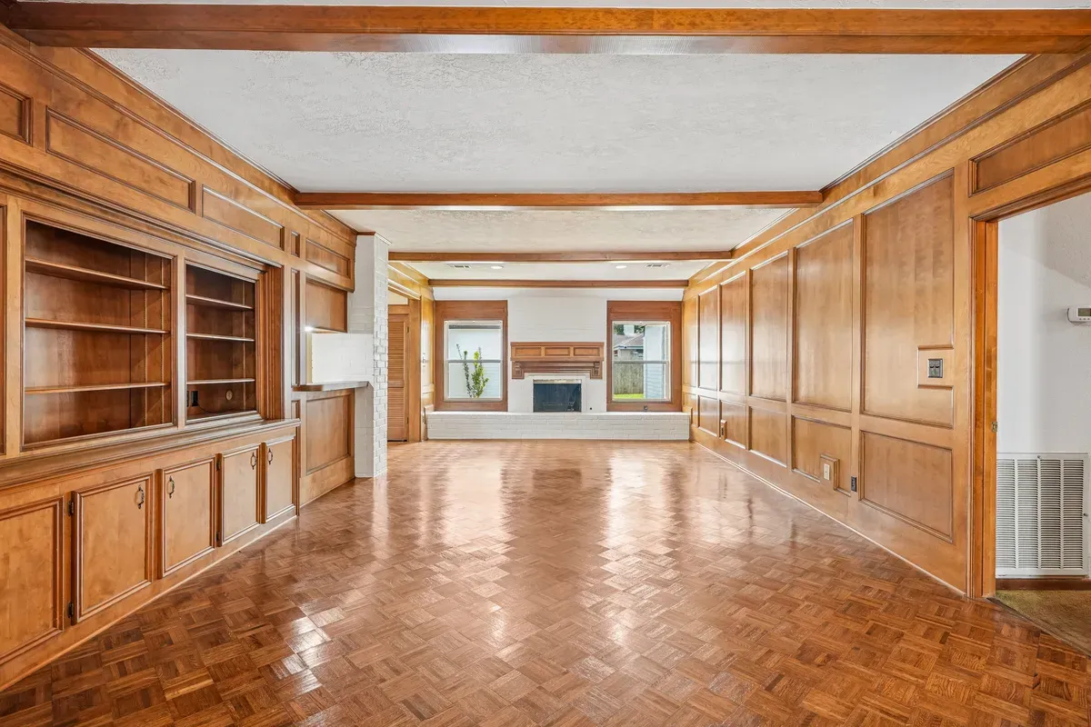 Empty wood-panelled living room before, with floor-to-ceiling timber panelling, built-in shelving, exposed beams, a white brick fireplace and a herringbone parquet floor