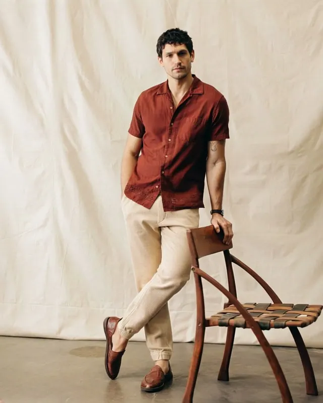 AI fashion video ad: a man in a rust short-sleeve shirt and linen trousers leaning on a wooden chair in front of a canvas backdrop